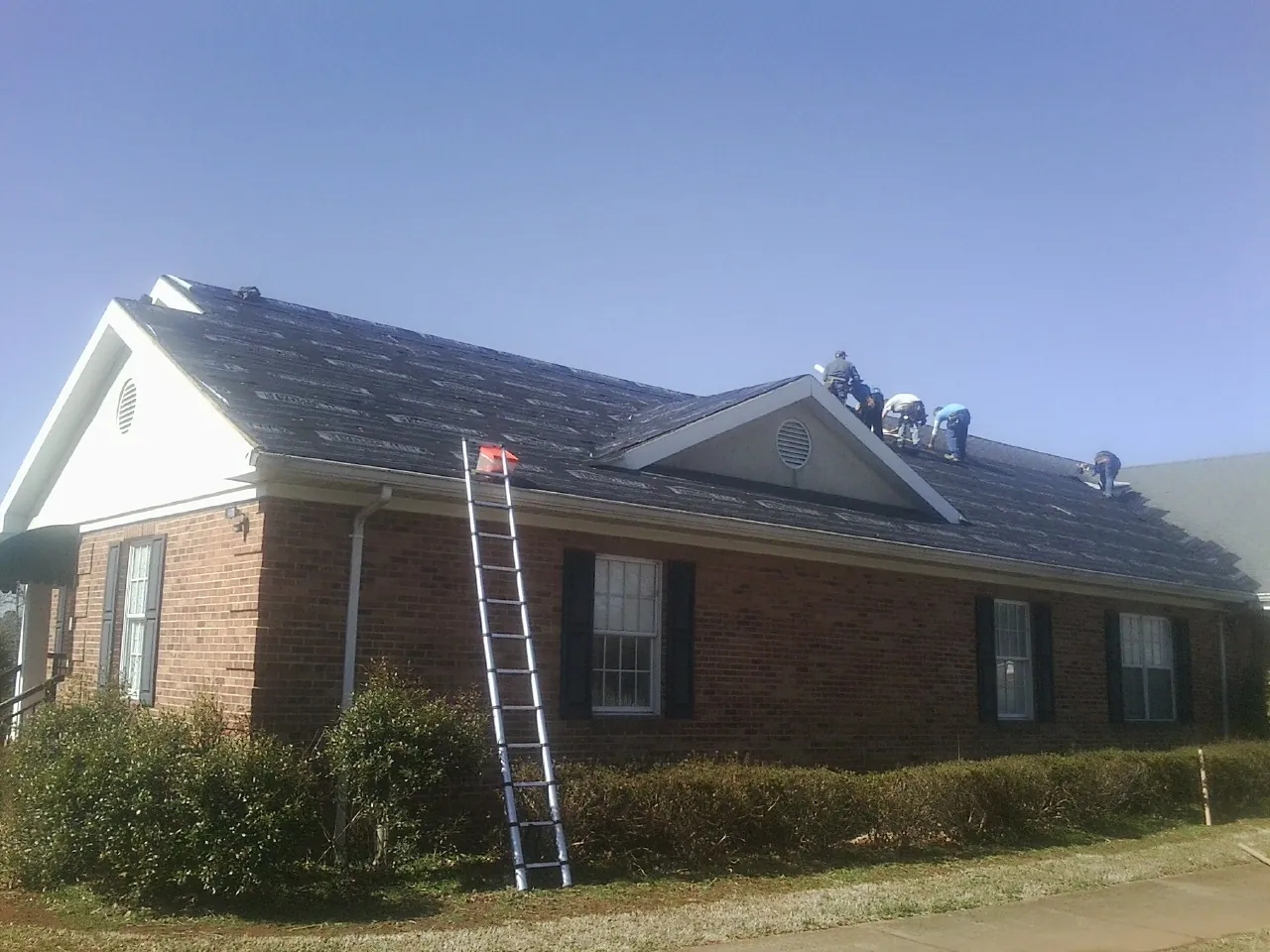 Crew tearing shingles off a brick ranch roof with a ladder set — Metal Roof Repair work by our crew in Vinita