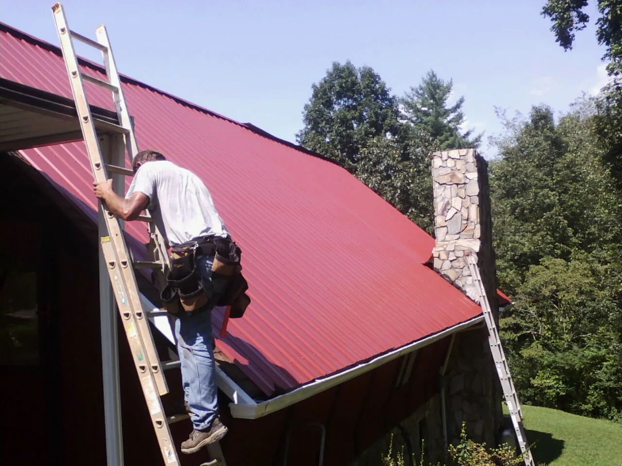 Roofer on a ladder beside a red metal roof and stone chimney — Soffit & Fascia Repair by Roofing Brothers in Vinita